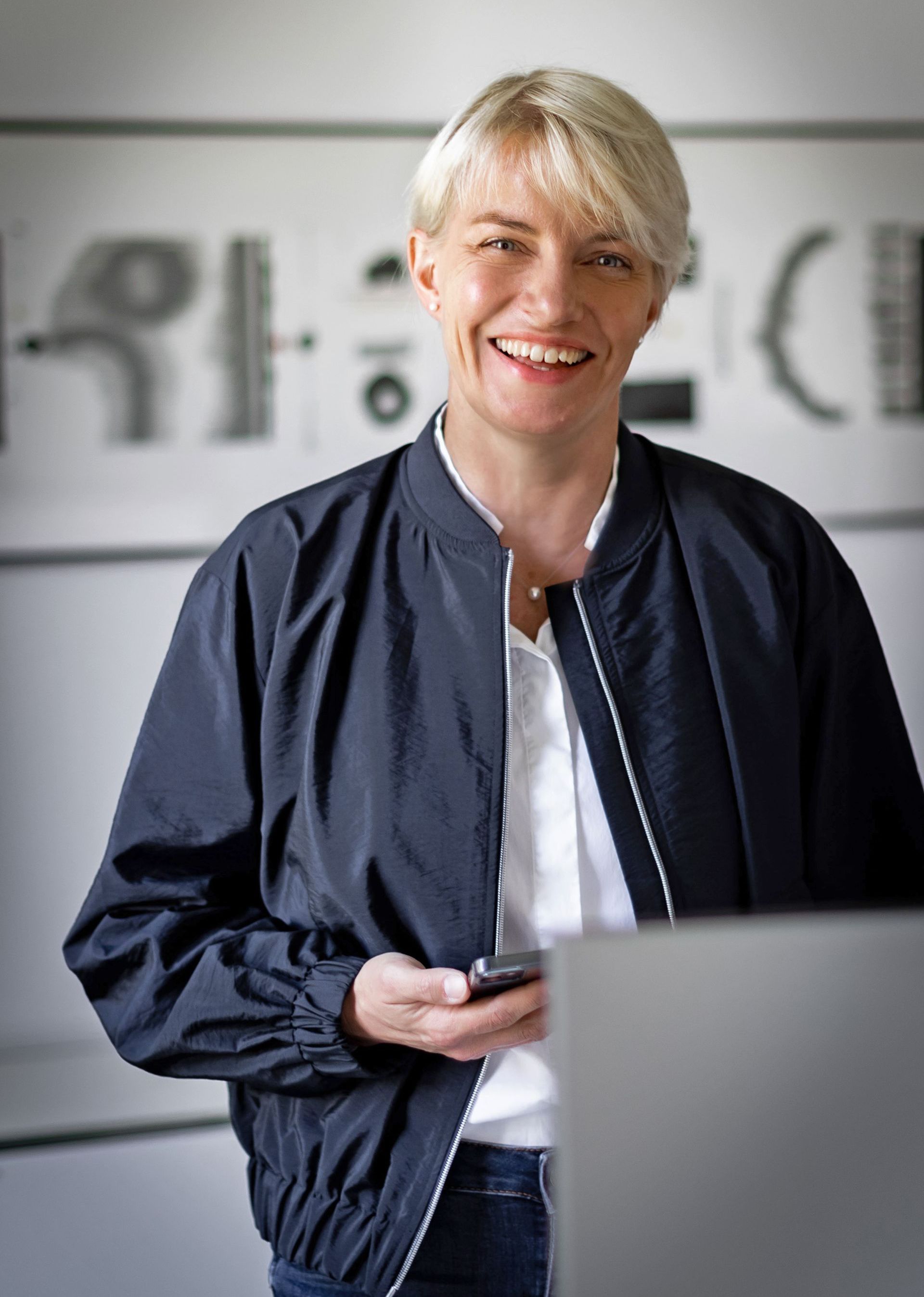 Eine Frau in einer schwarzen Jacke lächelt und hält ein Mobiltelefon