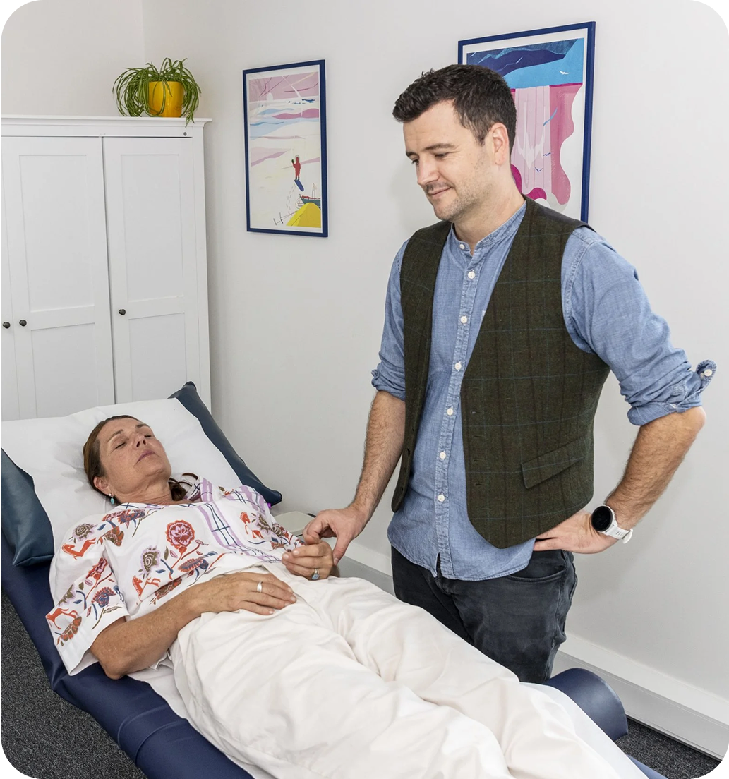 A female patient lies down, with her practitioner standing to her right, feeling her pulse.