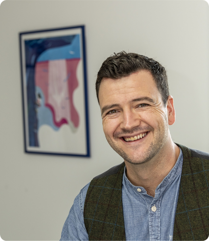 Josh Barrow smiling, leaning forward, wearing a blue shirt and green vest, with colourful abstract art in the background.