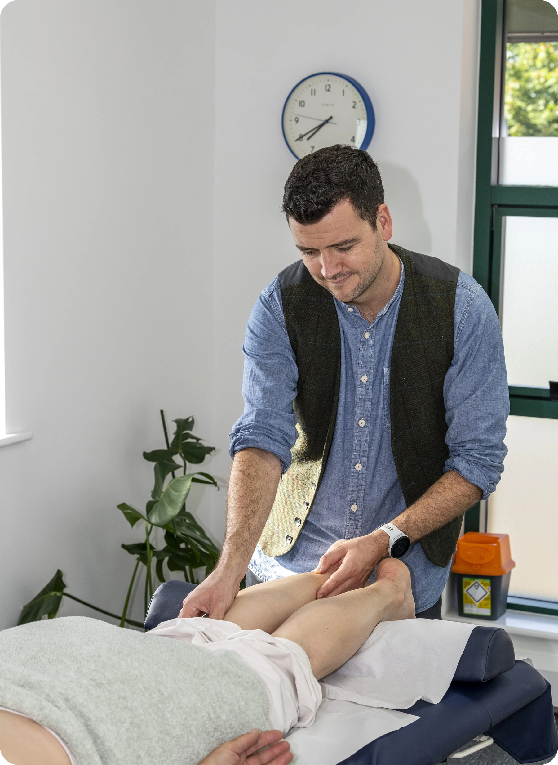 Man in vest examining a person's legs on a massage table in a treatment room.