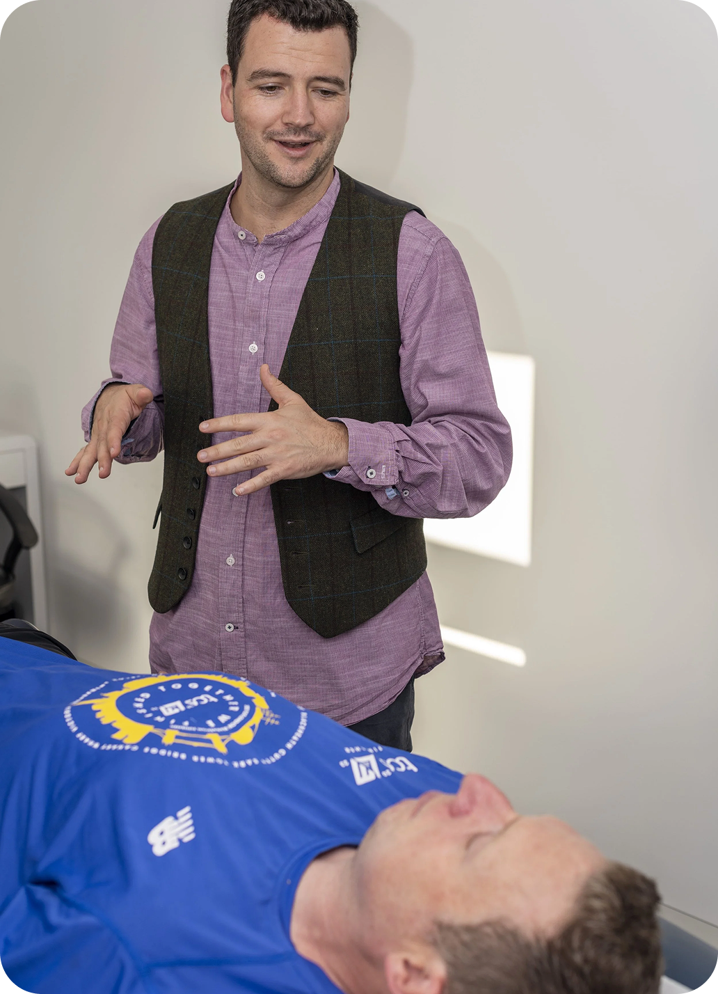 The acupuncturist, stood above a patient, is talking to the patient with his arms raised in an expressive and friendly way.
