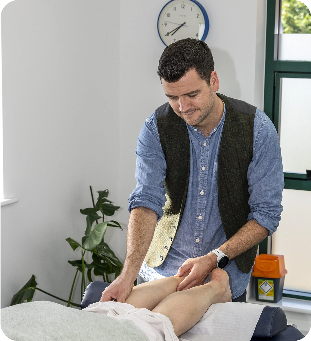 Man in vest massages patient's legs on a massage table. Room has a clock and a plant.