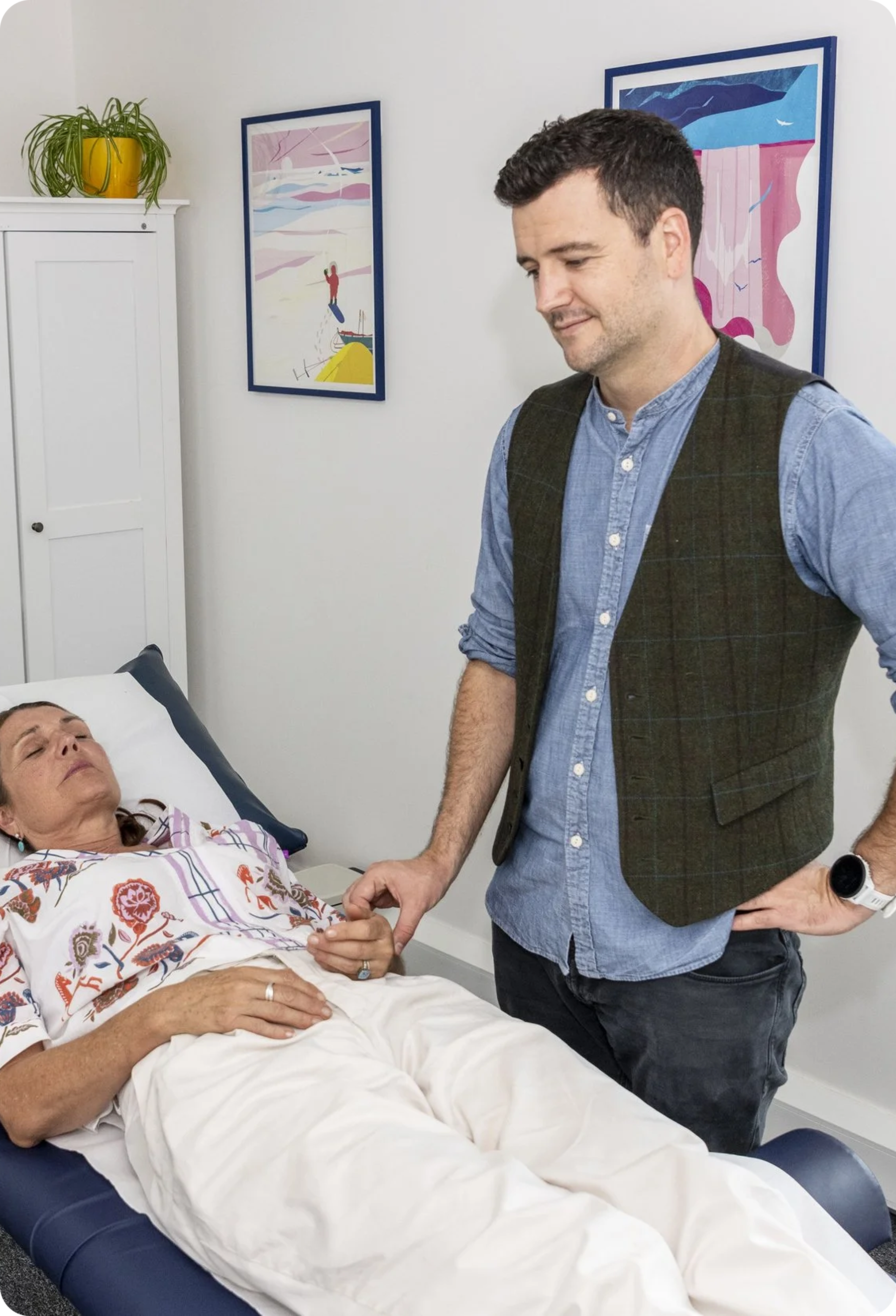 Man in vest standing over woman lying on medical table, holding her hand. Indoor setting, medical.