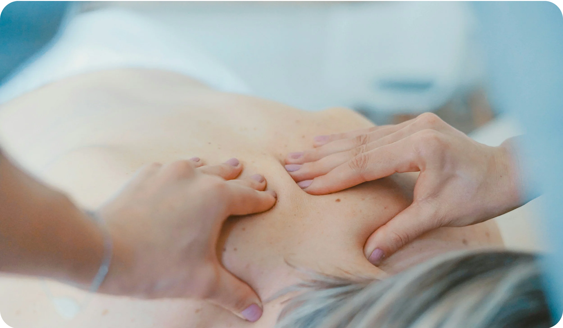 A patient is lying supine, with her back exposed, as a practitioner massages her upper back.