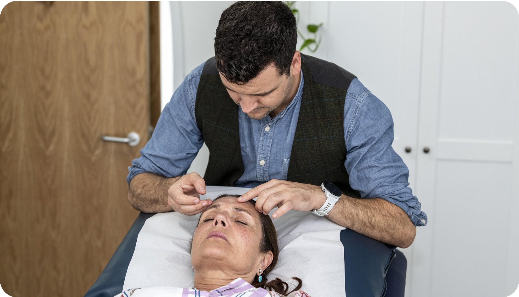 Man performing procedure on a person's forehead, possibly medical. In a room with wooden door.