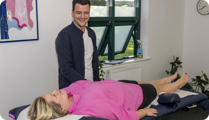 A smiling acupuncturist takes a patient's pulse as she lies on the couch.