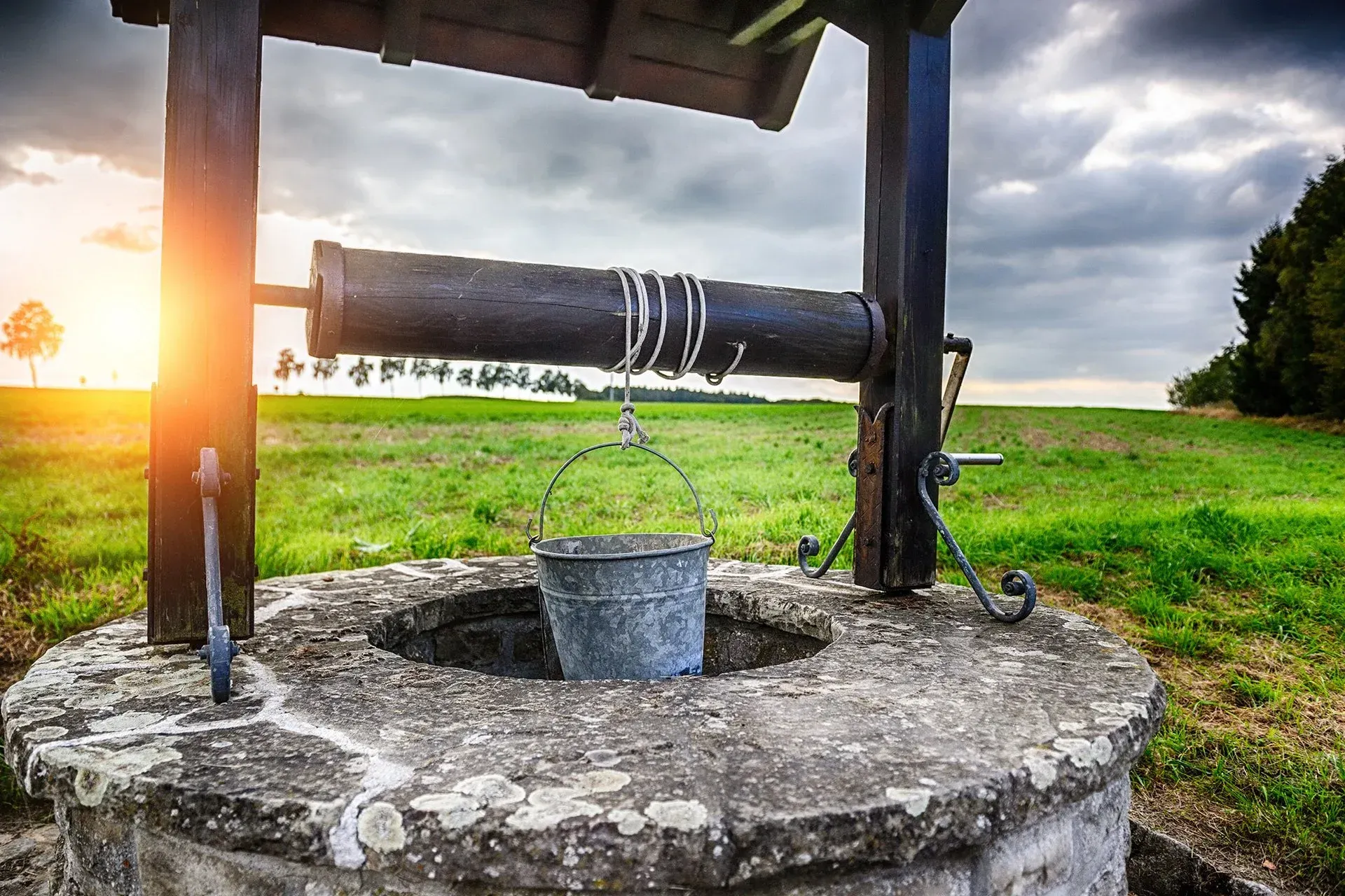 An image of a well in the foreground with some green grass behind and the sun rising.