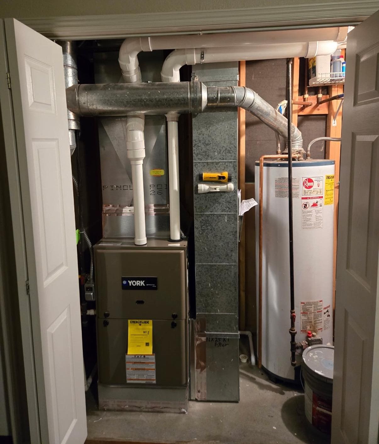 A utility closet containing a green gas furnace and a white Rheem water heater with overhead PVC piping.
