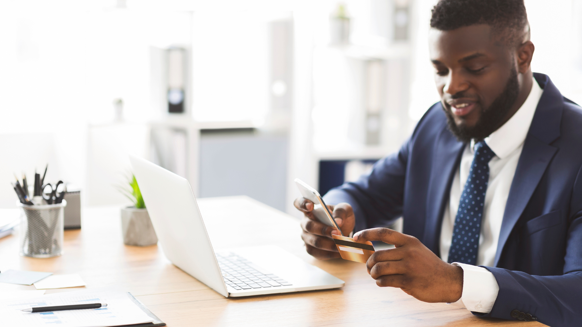 Man in suit smiling while using a smartphone at a desk with a laptop in a bright office
