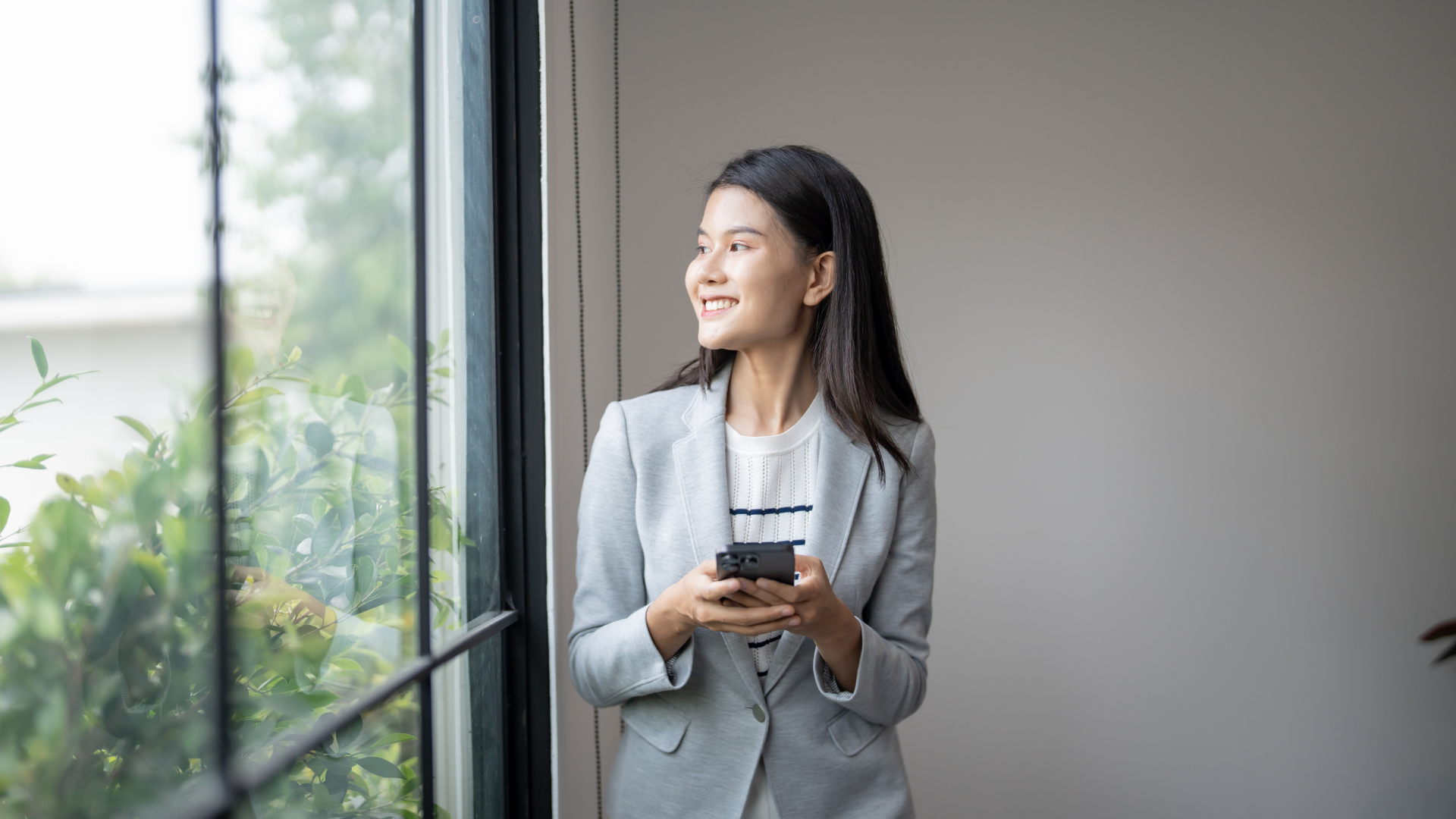 Woman in a light gray suit holding a phone, smiling by a window with greenery outside.