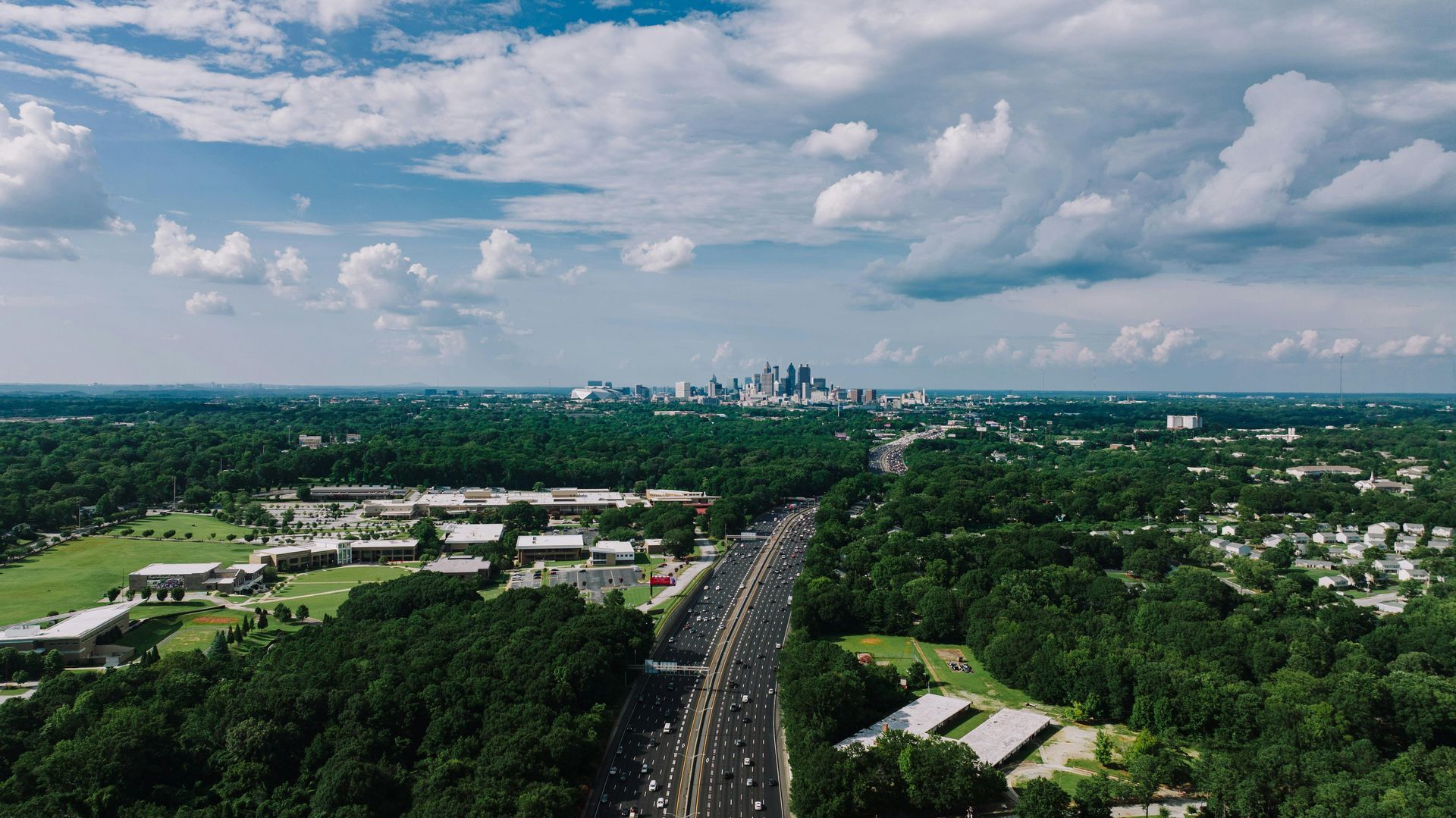 Aerial view of a highway cutting through green suburbs under a partly cloudy sky.