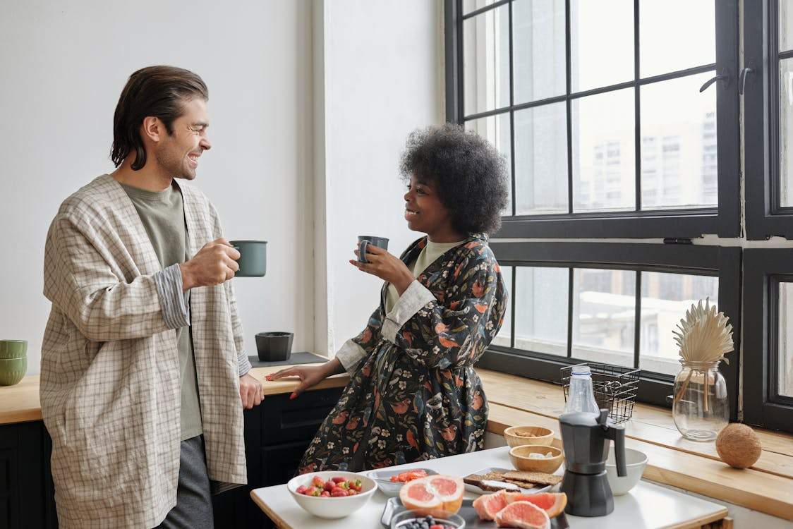 Two people chatting with mugs by a bright window, with a breakfast spread on the counter.