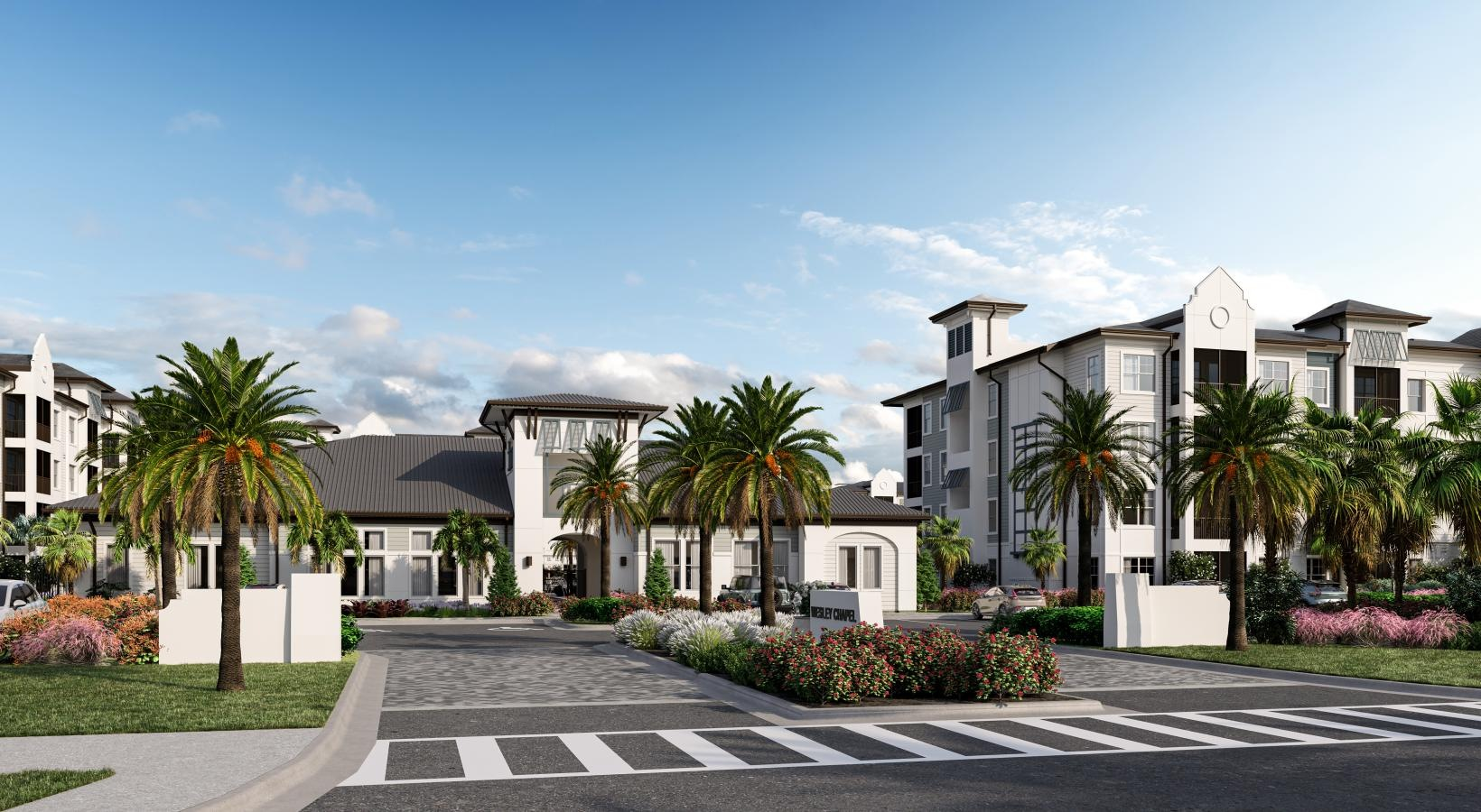 Modern apartment complex entrance with palm trees, landscaped gardens, and a crosswalk under a clear blue sky