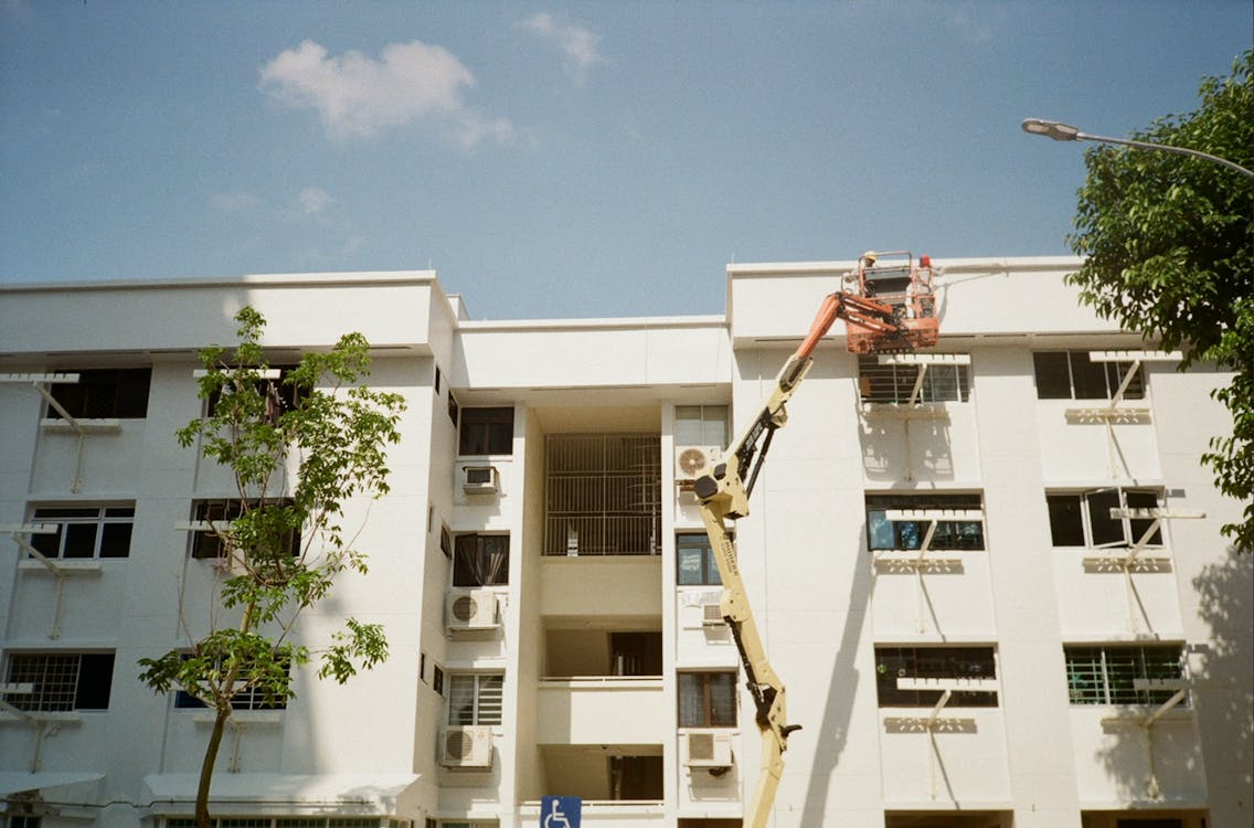 Worker on cherry picker trimming tree beside white apartment building under a clear blue sky