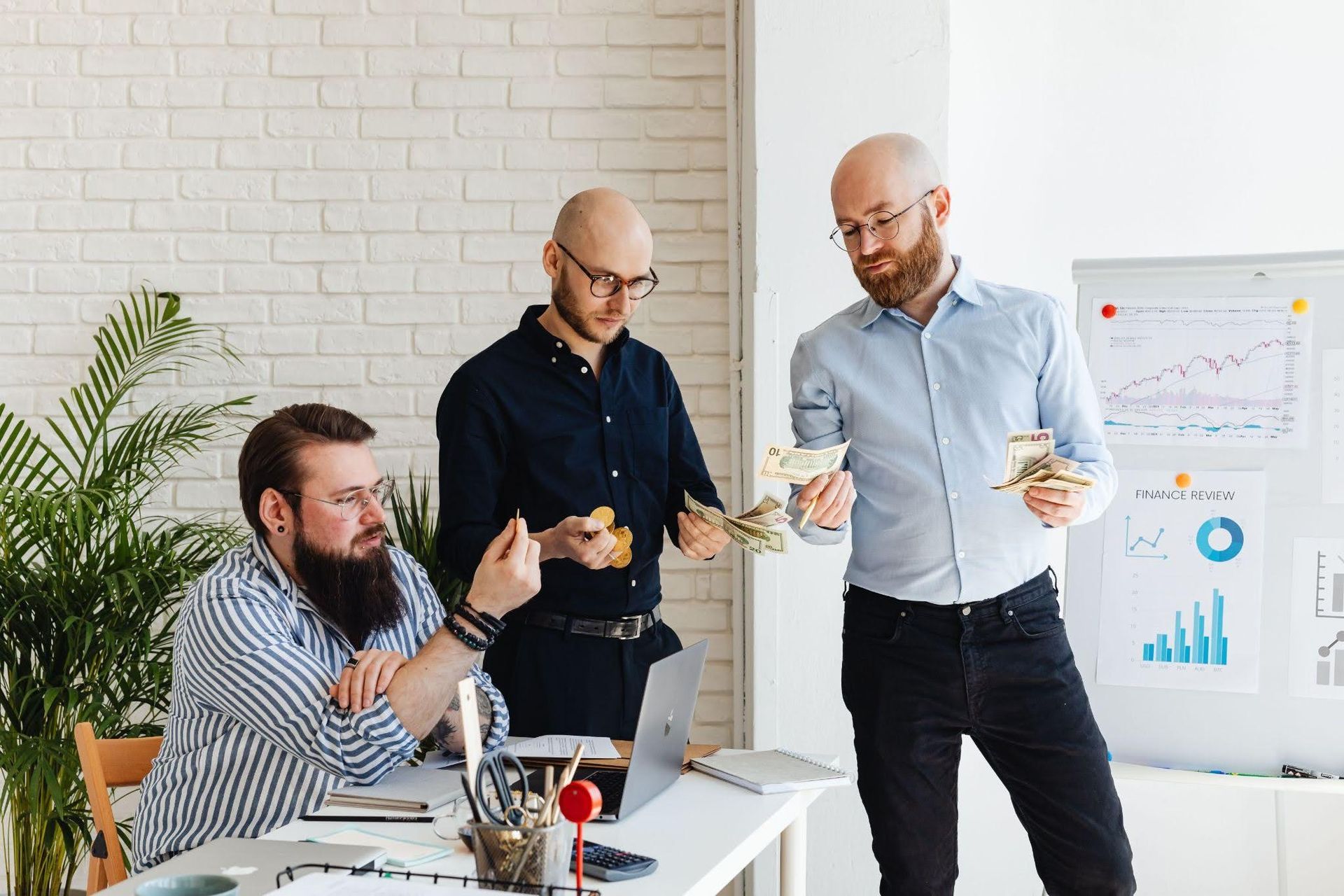 Colleagues discussing charts in a bright office, with one man seated and two standing by a presentation board.