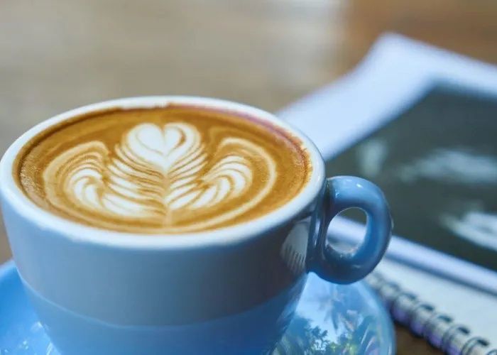 Blue cup of latte with heart-shaped foam art on a saucer beside a notebook