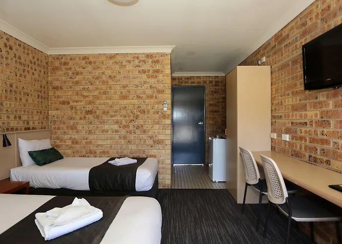 Small motel room with two beds, desk, TV, brick walls, and a mini fridge under a window.