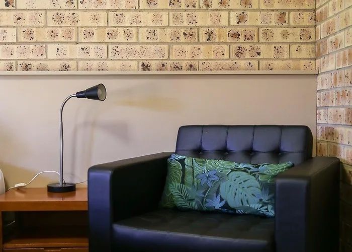 Black leather couch with green leaf pillows beside a floor lamp in a brick-walled room
