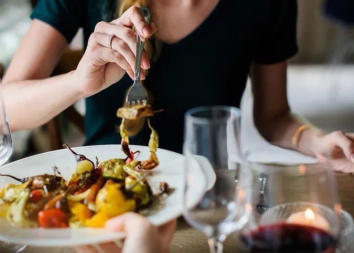 Person dining at a table, serving food from a plated meal with wine glasses nearby