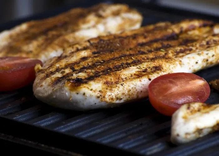 Grilled fish fillet with char marks cooking on a grill beside tomato halves and a lemon wedge