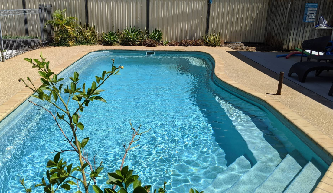 Backyard swimming pool with clear blue water, beige deck, and lounge chairs by a wooden fence.