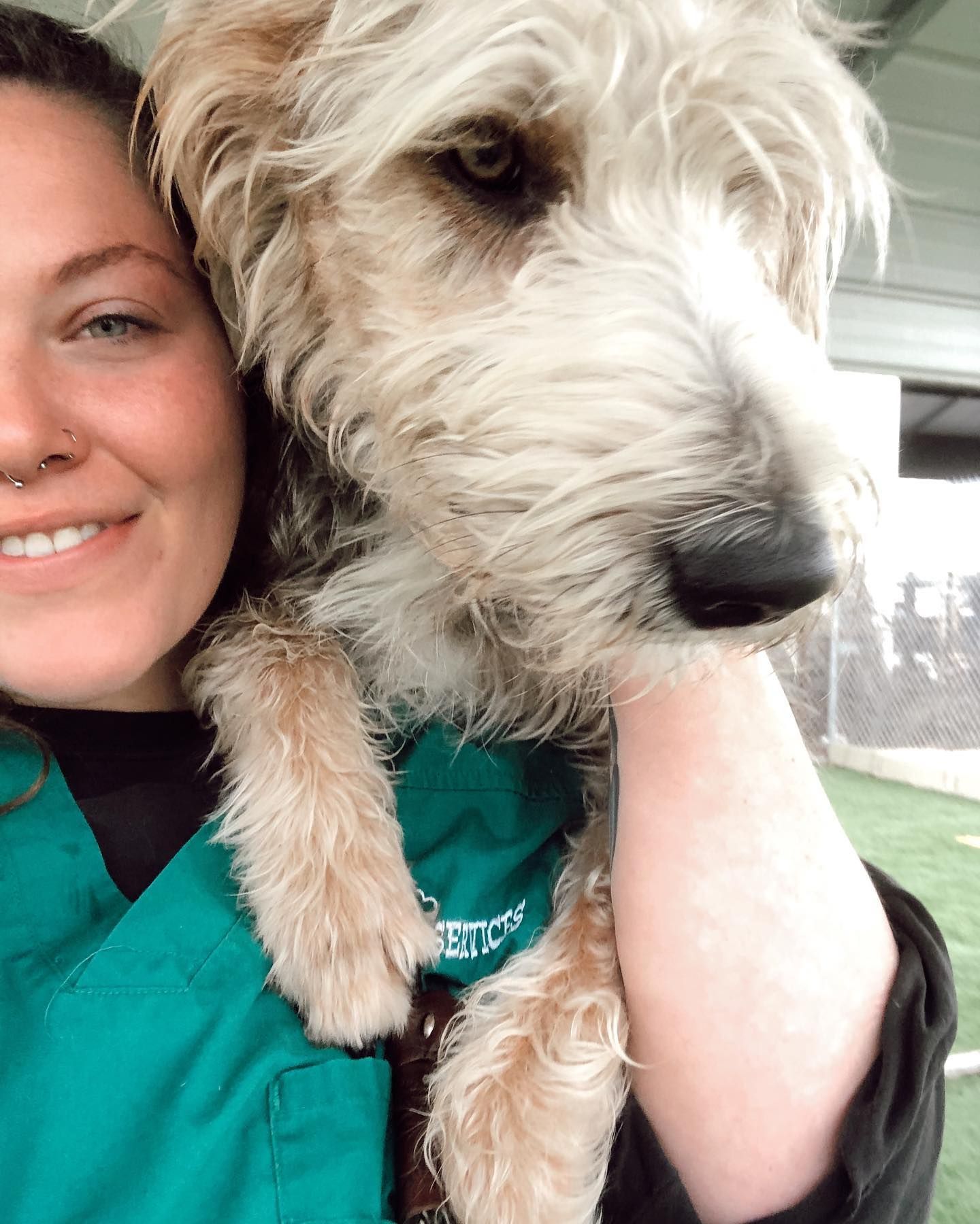 Woman holding a small, scruffy dog outdoors; both smiling at the camera. Dog wearing a green garment.