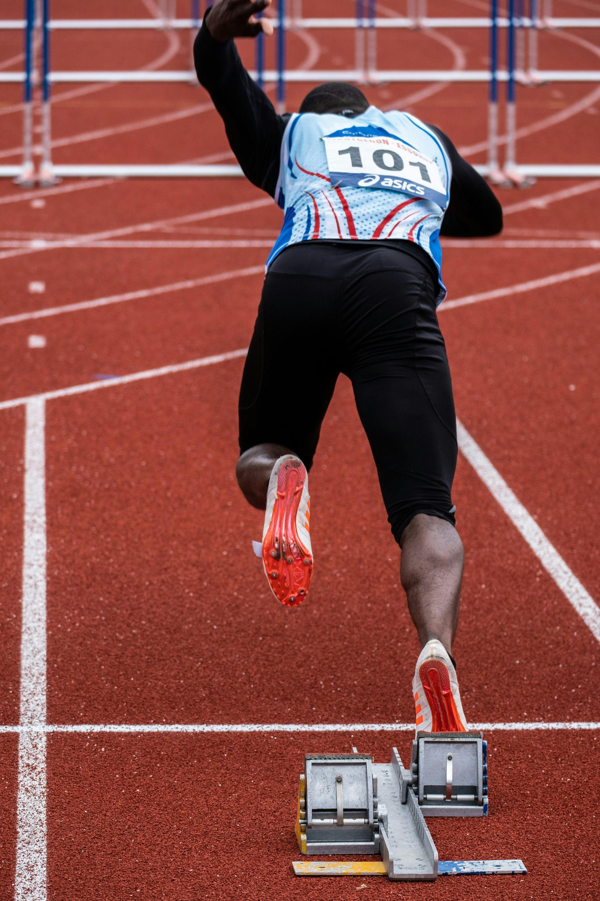 Track athlete sprinting from the starting blocks on a red running track, seen from behind.