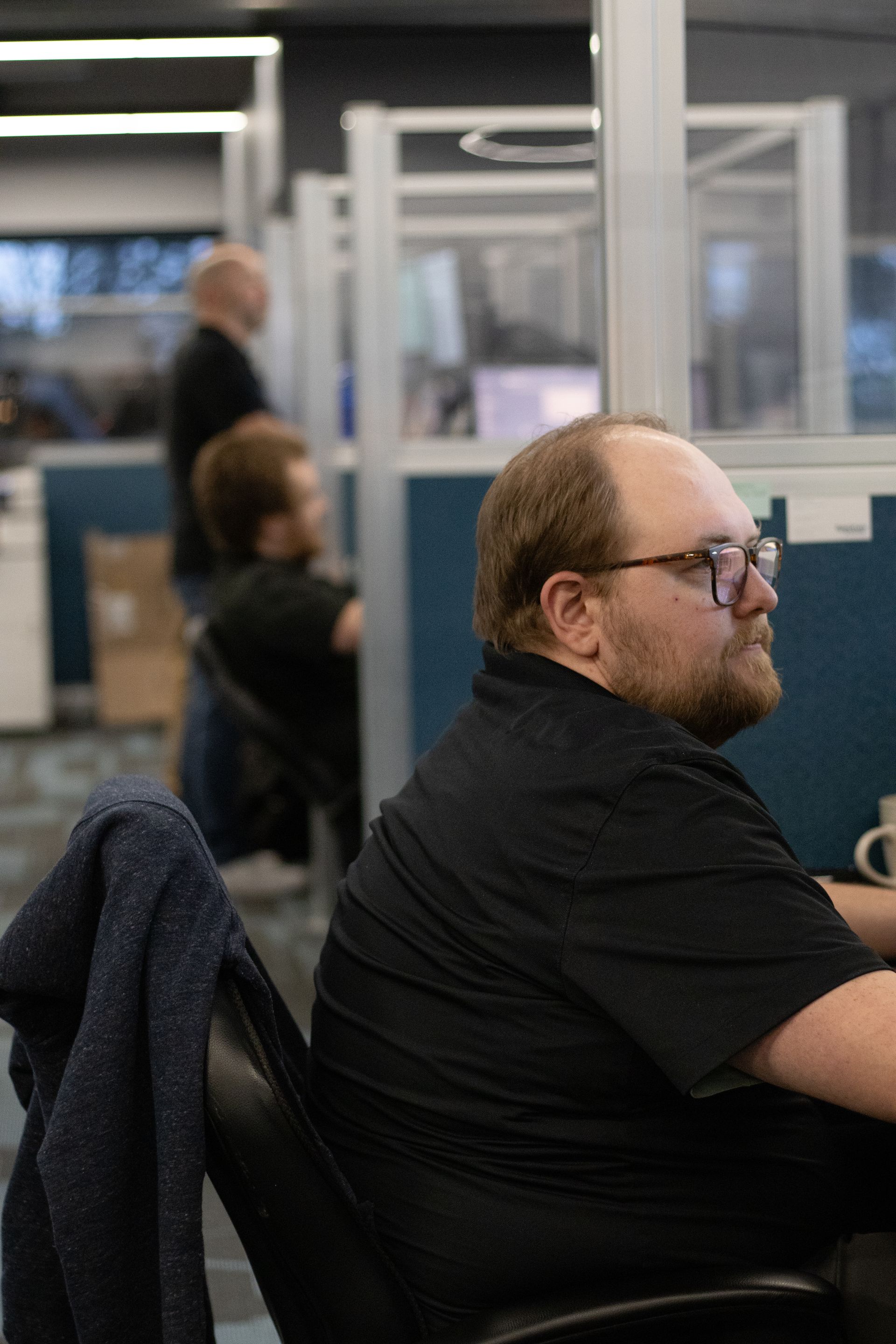 Man seated at a desk in an office, wearing glasses and a black shirt, with blurred coworkers in the background.