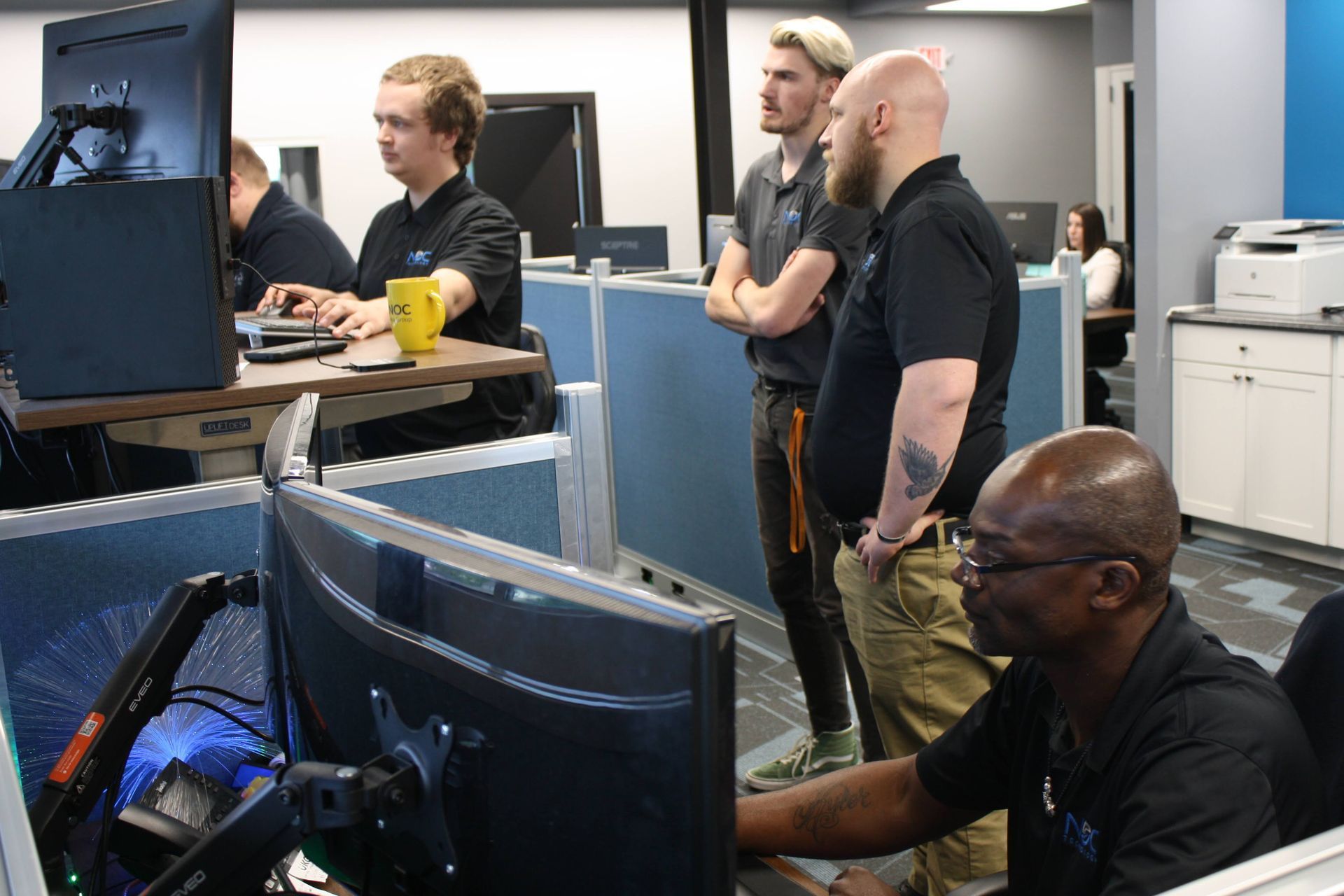 People working at computers in an office setting. One person is looking at a monitor, three others are observing.