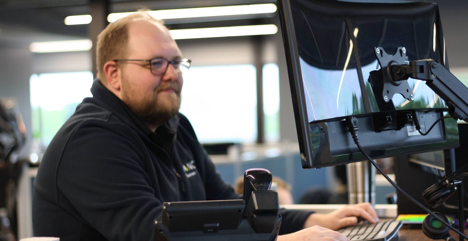 Man with glasses working at a computer in an office, smiling while looking at the screen and typing.