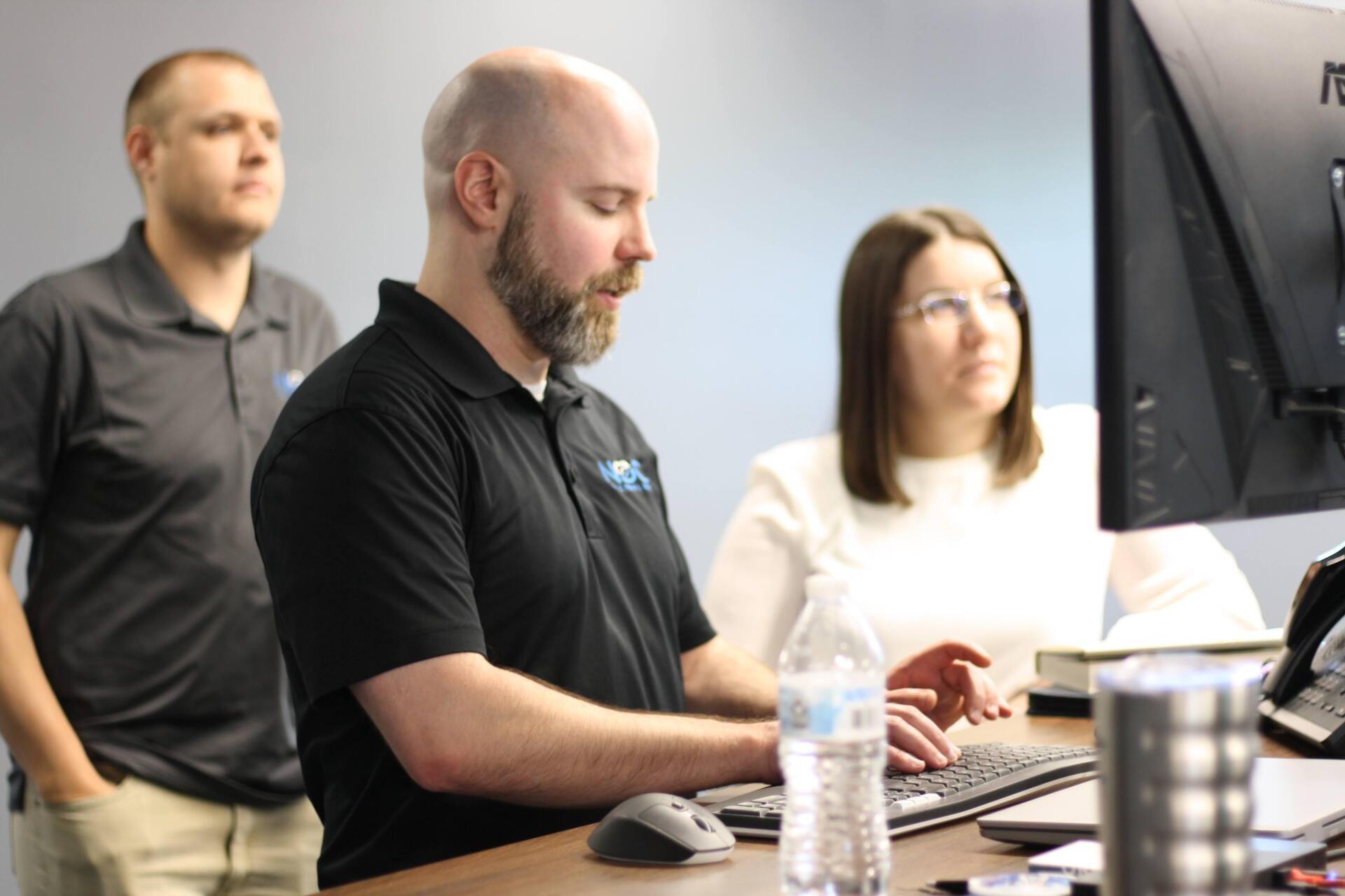 Three people in an office: one man types at a computer, another man looks on, a woman watches.
