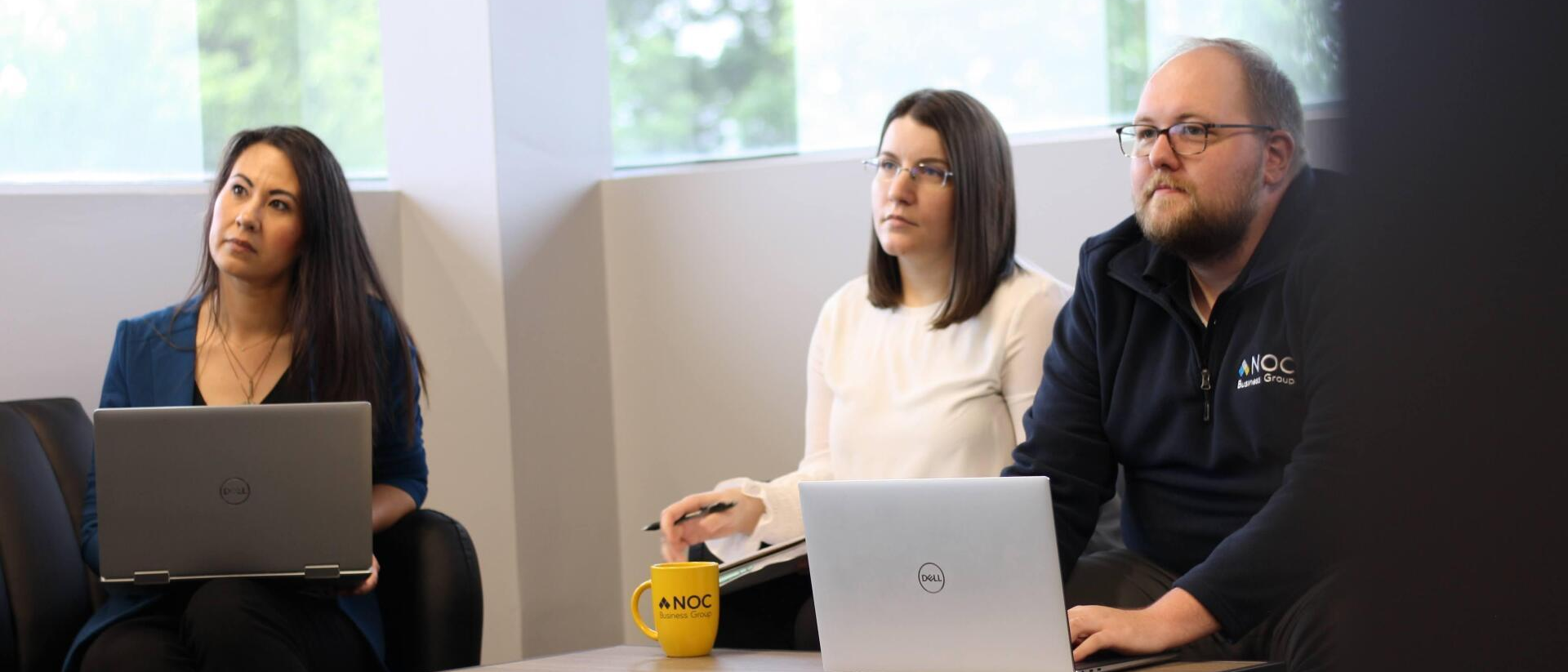 Three people in a meeting room looking up. Two women and a man. Laptops and a yellow mug are on the table.