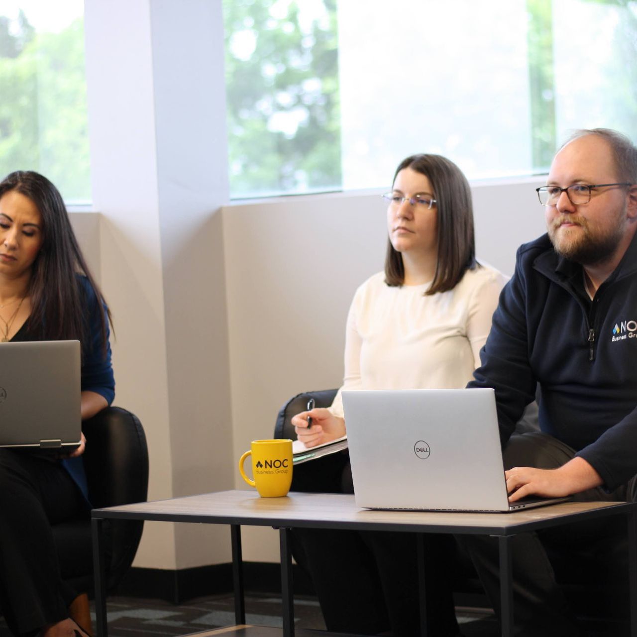 A group of people are sitting around a table with laptops and a yellow mug that says ' noc ' on it