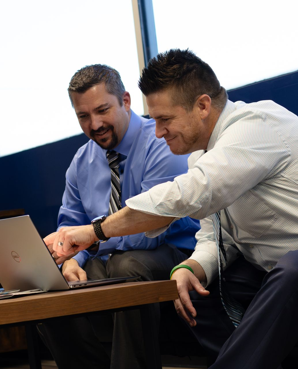 Two men in dress shirts and ties point at a laptop while seated at a table in an office