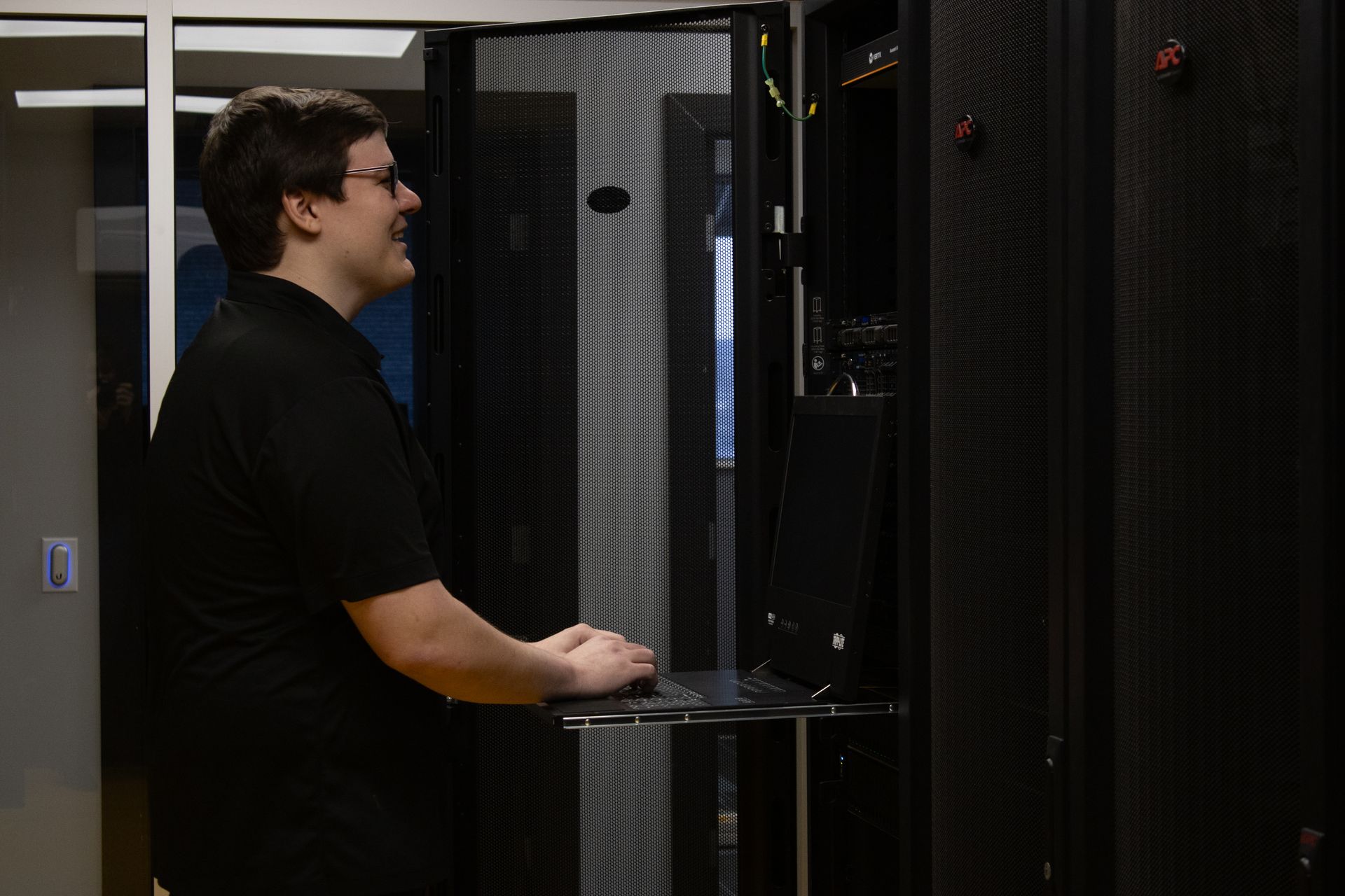 Person using a laptop in a server room, standing beside black server racks.