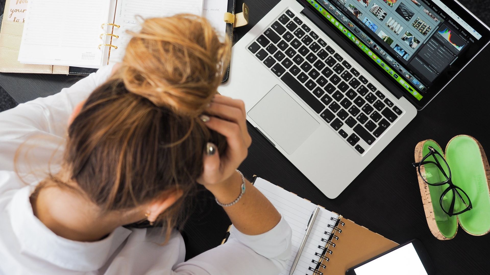 Woman with head in hands, overwhelmed at a desk with a laptop, notepad, and glasses needs IT support