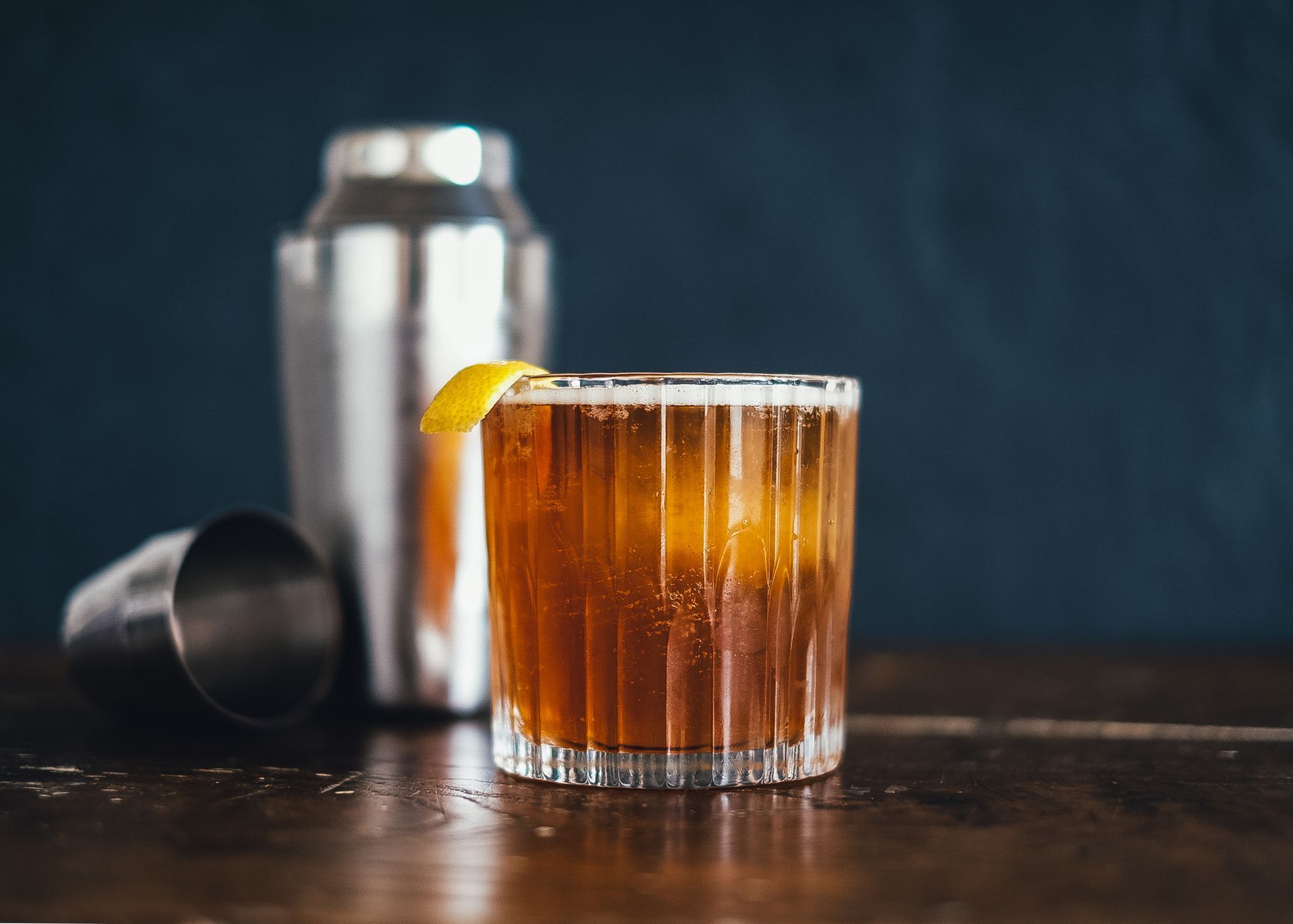 A glass of whiskey with ice and a lemon slice on a wooden table next to a shaker.