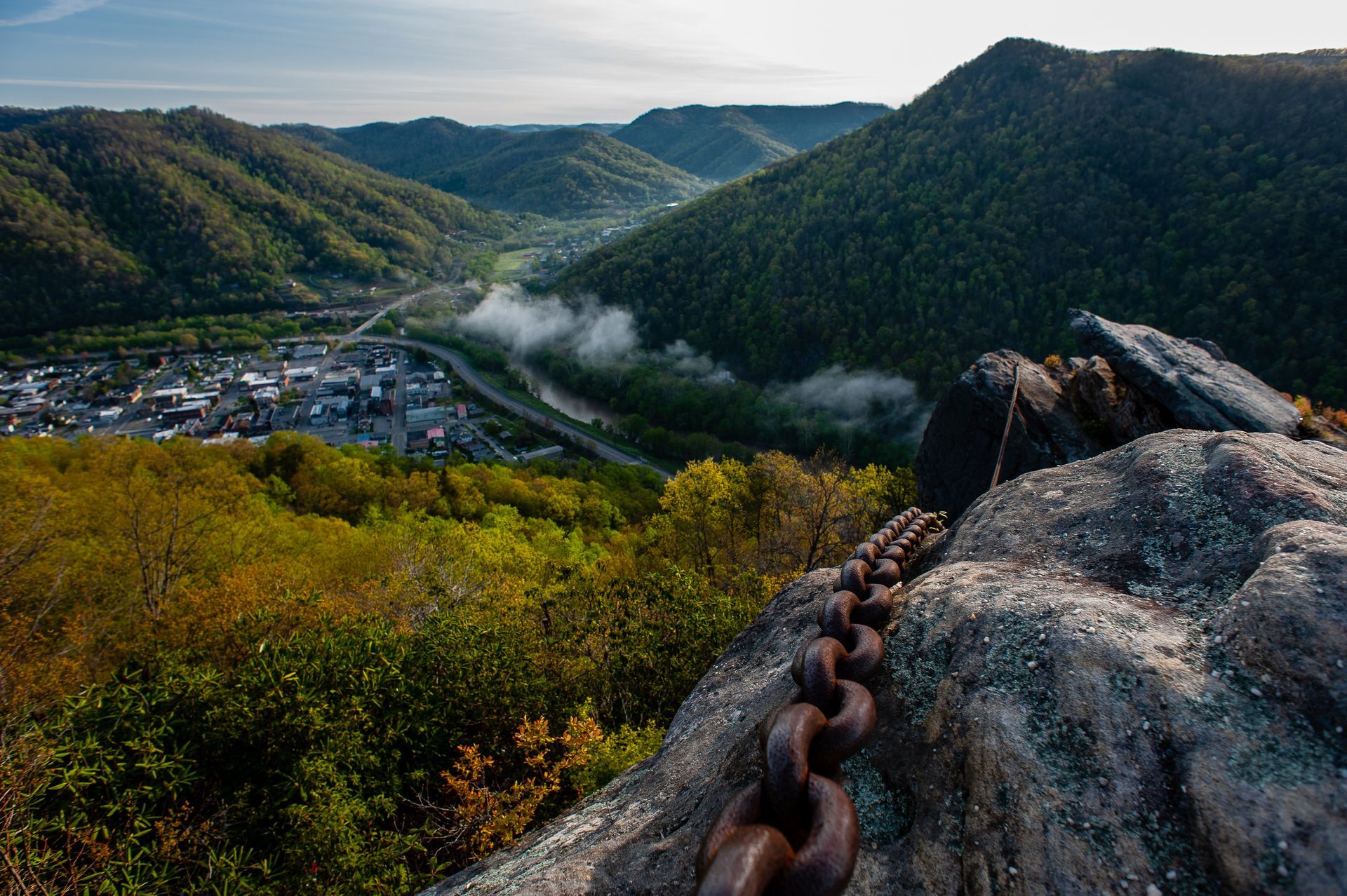 Overlooking a valley town, lush green mountains surround. Old chain lies on a rock in foreground.
