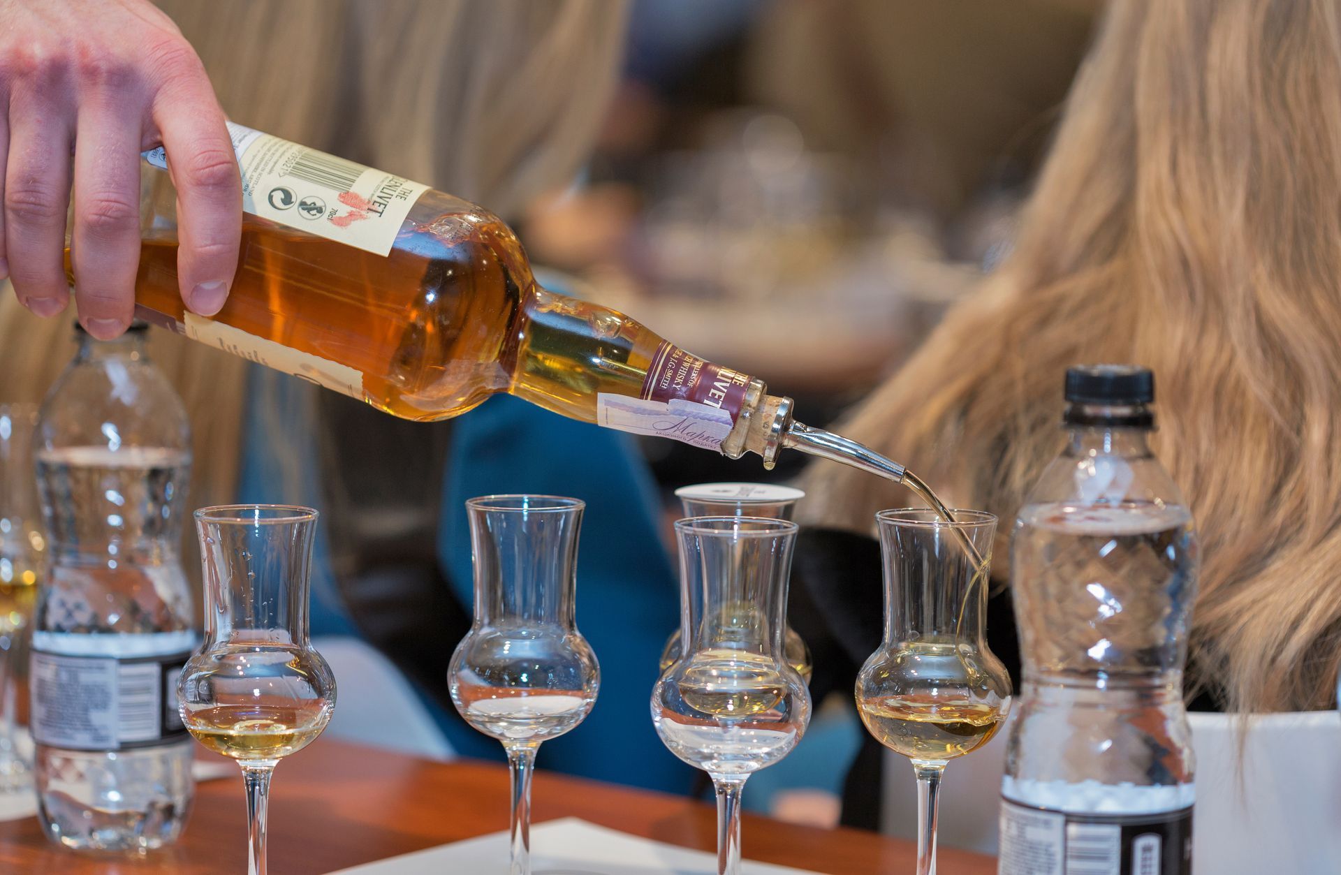 Someone pouring amber liquid from a bottle into small glasses, next to water bottles, at a table.
