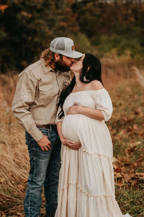 A man is kissing a pregnant woman on the cheek in a field.