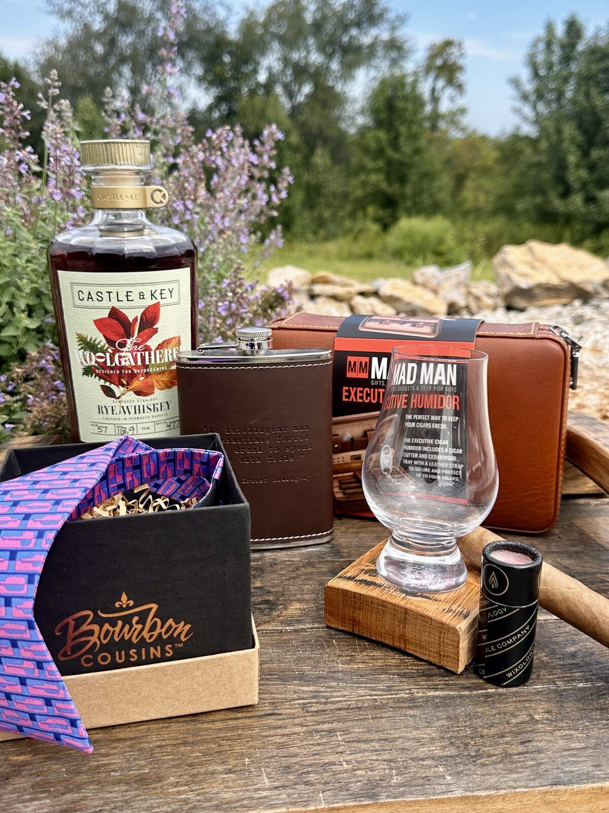 Navy and pink bourbon bow tie, leather flask, cigar humidor set, and Glencairn glass styled on a picnic table