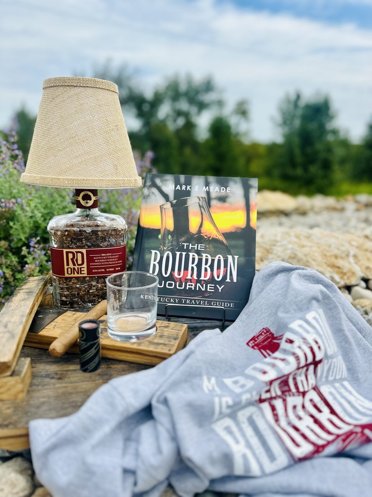 RD One bourbon bottle lamp, The Bourbon Journey Kentucky travel guide, rocks glass, and a bourbon t-shirt styled outdoors