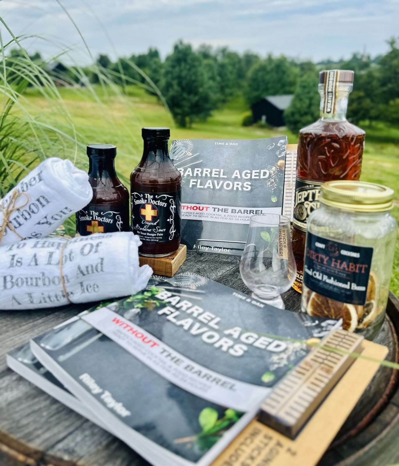 Barrel Aged Flavors cookbook, Smoke Doctors barbecue sauce, Glencairn glass, and bourbon bar towels styled together