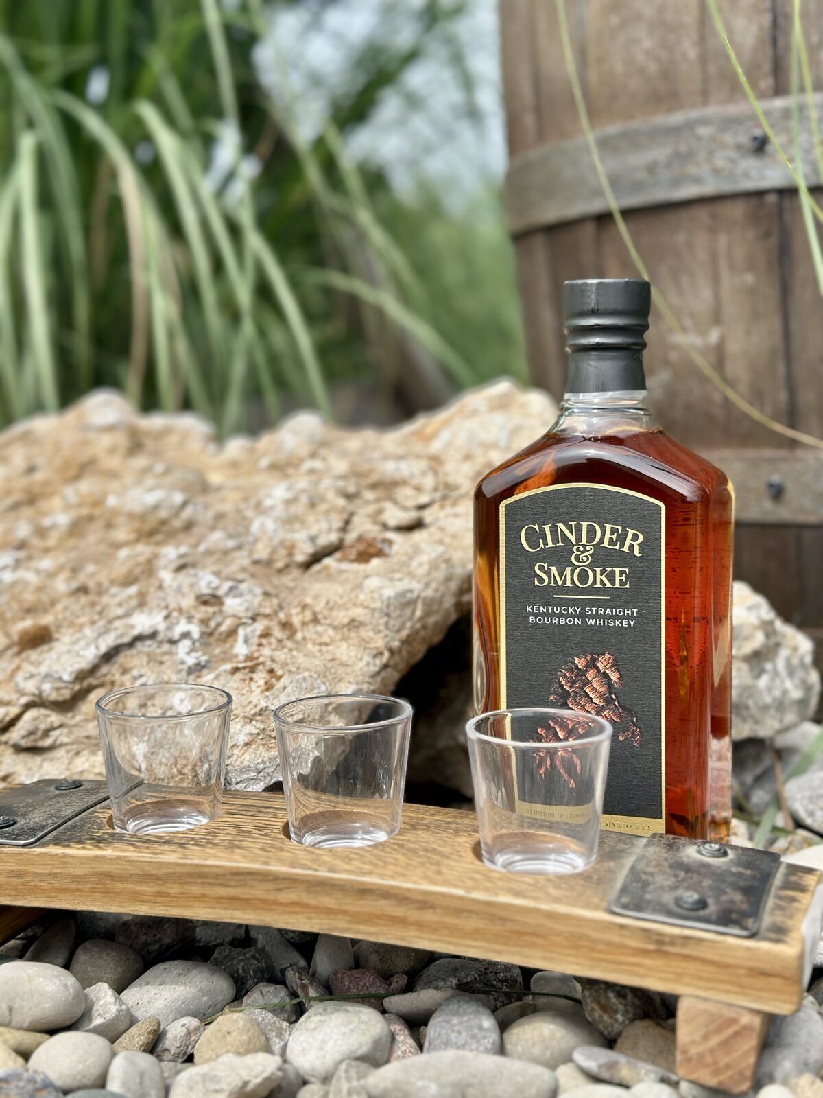 Raised three-glass bourbon stave flight board with tasting glasses and a bottle of Cinder & Smoke bourbon