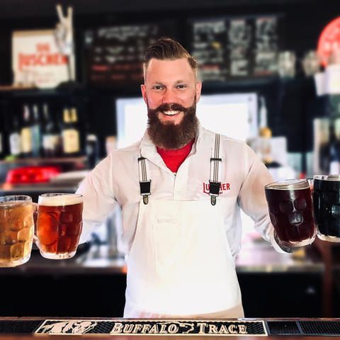 A man with a beard is standing behind a bar holding three mugs of beer.