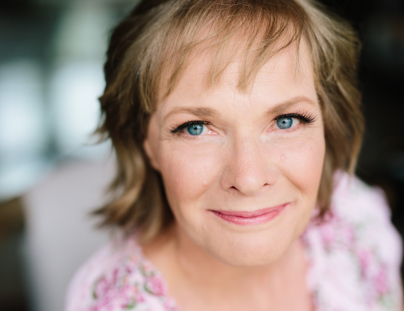 Smiling woman with blue eyes and short hair in a pink floral top, softly blurred background