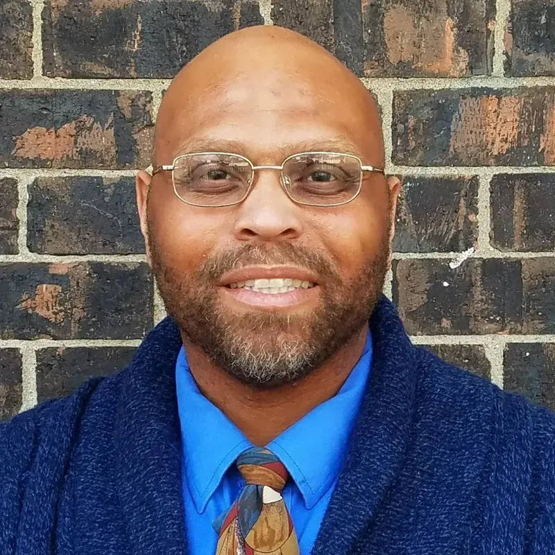 Smiling bald man in glasses, blue shirt and patterned tie, standing before a brick wall.