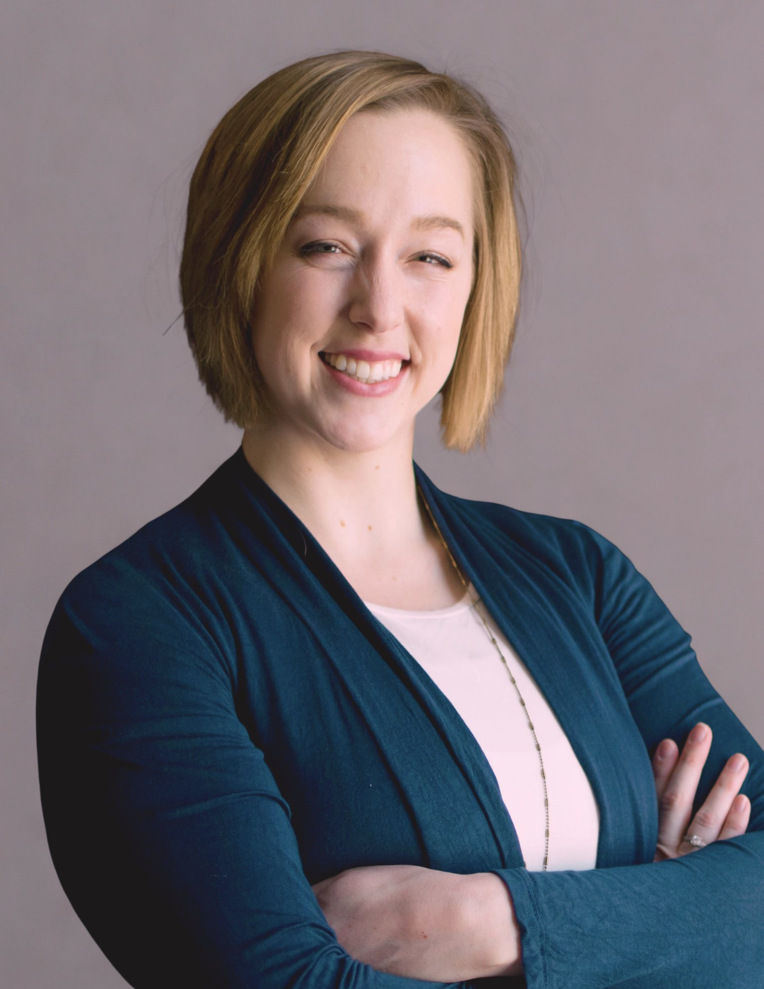 Smiling woman with short blonde hair, arms crossed, wearing a teal cardigan against a gray background