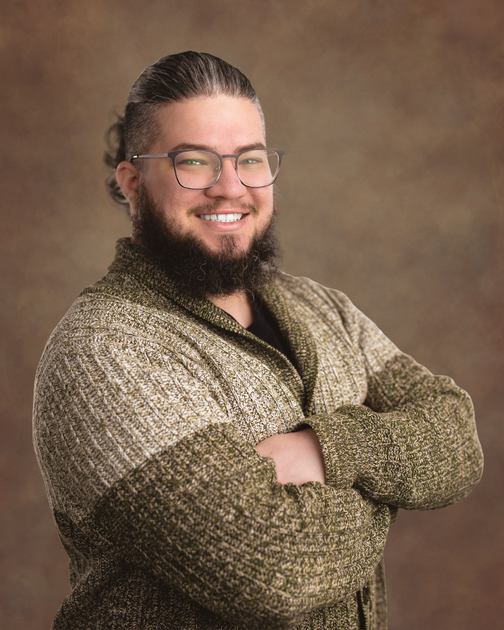 Caleb Duron, Smiling person with glasses and a beard, arms crossed, wearing a textured sweater against a brown backdrop
