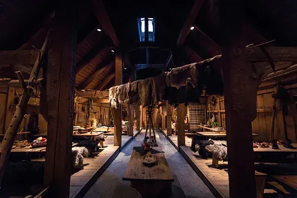 Dim rustic dining hall with long tables, hanging clothes, and warm lantern light.